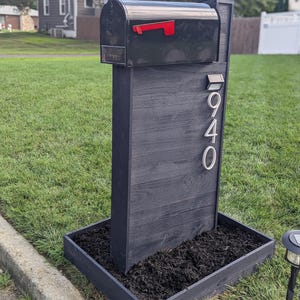 May include: A black mailbox with a red flag, mounted on a dark grey wooden post. The post features silver house numbers and sits in a black planter box filled with mulch. The mailbox is in a grassy garden.