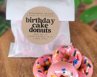 May include: Three pink cake donuts with colorful sprinkles. The donuts are packaged in a white bag with a brown label that reads "Weekend Wicks Candle Company birthday cake donuts Wax Melts Hand Poured in FL 2.00Z (57G)"