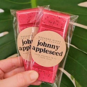 K&ouml;nnte beinhalten: Zwei rote Wachsschmelzen, verpackt in durchsichtigen Plastikt&uuml;ten. Die Wachsschmelzen sind mit "johnny appleseed" und "Weekend Wicks Candle Company" beschriftet.