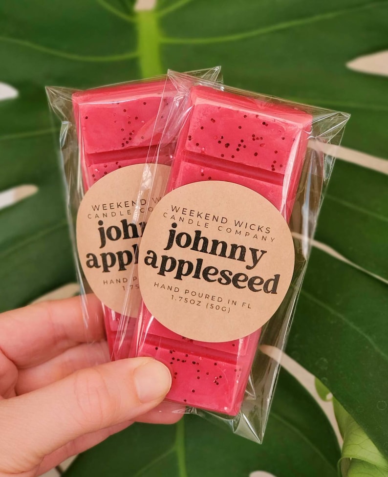 K&ouml;nnte beinhalten: Zwei rote Wachsschmelzen, verpackt in durchsichtigen Plastikt&uuml;ten. Die Wachsschmelzen sind mit "johnny appleseed" und "Weekend Wicks Candle Company" beschriftet.