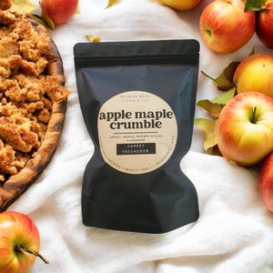 May include: A black bag of "Apple Maple Crumble" carpet freshener sits on a white surface, surrounded by red and yellow apples and fall leaves. The bag has a circular label with the product name and ingredients.