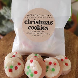 May include: Six round wax melts shaped like cookies with white frosting and red and green sprinkles. The wax melts are packaged in a white paper bag with a label that reads "Weekend Wicks Candle Company Christmas Cookies Wax Melts Hand Poured in FL 2 oz (57g)"
