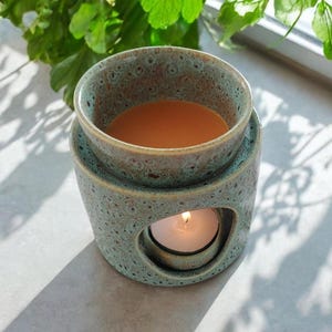 May include: A teal ceramic wax warmer with a speckled pattern. The warmer has a round top dish filled with a liquid, and a lower compartment with a lit candle. The design includes a circular opening for the candle's light to shine through.