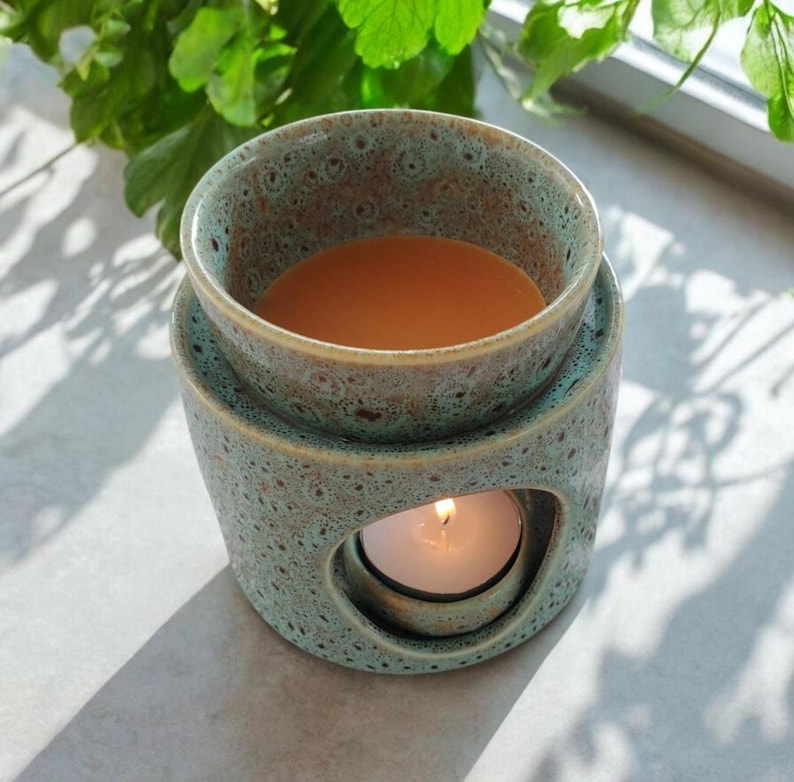 May include: A teal ceramic wax warmer with a speckled pattern. The warmer has a round top dish filled with a liquid, and a lower compartment with a lit candle. The design includes a circular opening for the candle's light to shine through.