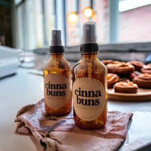 以下が含まれることがあります： 黒いスプレーキャップと「cinna buns」と黒文字で書かれたラベルが付いた、琥珀色のガラス瓶が2本。瓶は茶色の布地に置かれている。