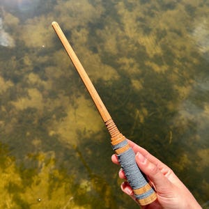 May include: A handcrafted wooden wand with a light brown finish. The handle is wrapped with gray thread and accented with thin, light brown bands. The wand is held against a blurred background of water, creating a natural and magical aesthetic.