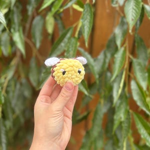 May include: A small, handmade, yellow and brown crocheted bee with white wings. The bee is held in a hand against a background of green leaves and a wooden fence. The bee is a cute, whimsical item.