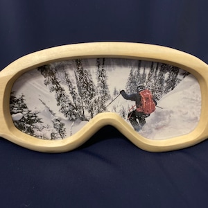 Peut inclure: Un cadre de lunettes de ski en bois avec une photo d'un skieur dans une forêt enneigée. La photo montre un skieur en veste rouge et sac à dos, descendant une pente enneigée.
