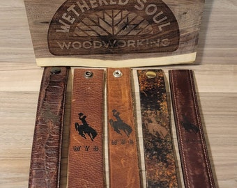 May include: A wooden sign with the text "WETHERED SOUL WOODWORKING" above five leather straps. The straps are various shades of brown and feature a "WYO" logo with a cowboy on a horse. Each strap has a metal snap.
