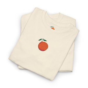 Peut inclure: Deux t-shirts crème superposés, chacun orné d'un motif brodé d'orange. L'orange est un mélange vibrant de teintes rouges et oranges, avec des feuilles vertes. Le motif est répété sur le haut du côté gauche de la poitrine des chemises.