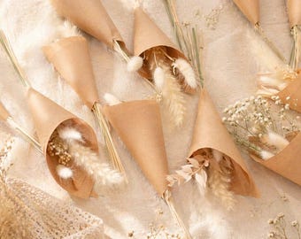 May include: An overhead shot of several small bouquets of dried flowers wrapped in brown paper cones. The flowers are a mix of beige, cream, and white, with feathery textures. The cones are arranged on a light-colored, textured surface.