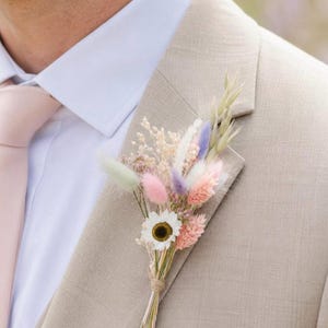 Op de afbeelding: Beige colbert met een bloemcorsage in pastelkleuren. De corsage heeft een witte madelief, roze, paarse en groene gedroogde bloemen, vastgebonden met touw. Een lichtroze stropdas en een wit overhemd met kraag zijn ook zichtbaar.
