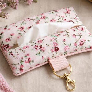 May include: A floral print tissue holder in pink and burgundy. The rectangular holder has a pink border, a pink strap, and a gold-colored clasp. White tissues are visible in the opening.