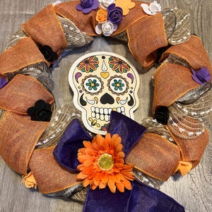 May include: A decorative wreath featuring a Day of the Dead sugar skull centerpiece. The wreath is made of orange burlap ribbon, accented with purple, orange, and white flowers, and a large purple bow with an orange flower.