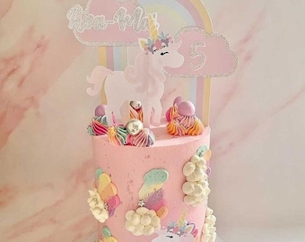 May include: A pink frosted cake decorated with a unicorn theme. The cake features a unicorn topper, rainbow accents, and the number 5. Colorful frosting and decorative elements adorn the sides and top of the cake, creating a festive look.