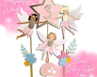 May include: A whimsical cake topper featuring three fairy figures with glittery gold accents, pink dresses, and white wings. A pink star with the number 3 is at the top. The text reads "Personalise Wand" and "Choose Gold or Silver Glitter".