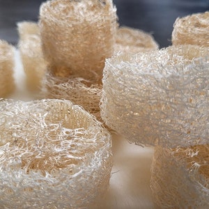 May include: A collection of cylindrical, beige loofah sponges. The natural, fibrous texture of the sponges is visible, with a light color palette. These bath accessories are arranged in a close-up shot.