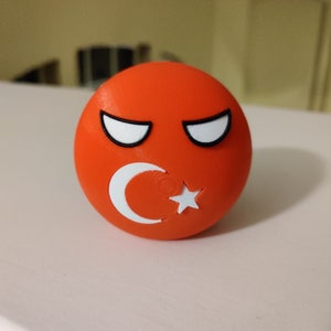 May include: A red 3D printed sphere with a white crescent moon and star, representing the Turkish flag. The sphere has a cartoon-like face with black eyebrows and white eyes.