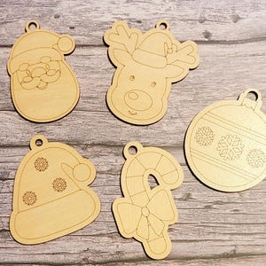 May include: Five unfinished wooden Christmas ornaments. The ornaments are shaped like Santa Claus, a reindeer, a Christmas tree ornament, a Santa hat, and a candy cane. The ornaments are ready to be painted.