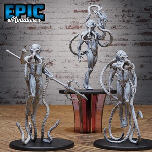 May include: Three grey alien miniature figures with tentacle-like appendages. One figure holds a trident, another stands on a table, and the third stands on a black base. The figures are detailed and stylized.