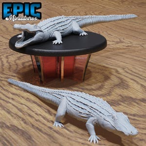 May include: Two light gray unpainted crocodile figurines with textured scales. One crocodile is positioned on a black circular pedestal with a translucent red base, while the other rests on the surface. The text Epic Miniatures is visible.