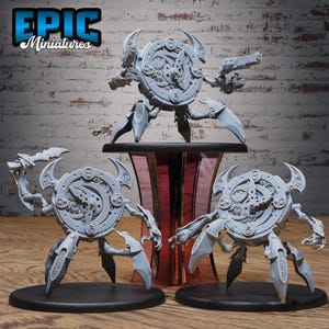 May include: Three gray miniature robot figures with clockwork-inspired designs. Each figure has a circular body with gears, horns, and articulated limbs. One figure is on a red and black stand, while the others are on black bases. The text "EPIC Miniatures" is in the upper left corner.