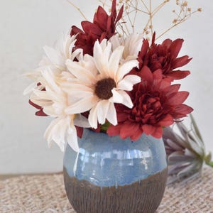 May include: A blue and brown ceramic vase holds a bouquet of artificial flowers. The flowers are a mix of white, cream, and burgundy. The vase has a textured, dark brown base and a smooth, light blue top.