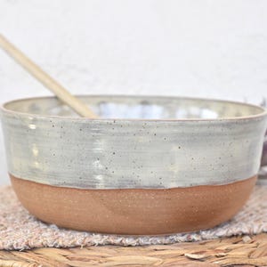 May include: A handmade ceramic bowl with a two-tone design. The upper portion is a speckled off-white, while the lower half is a natural terracotta color. A wooden spoon rests inside the bowl, which is placed on a woven mat.
