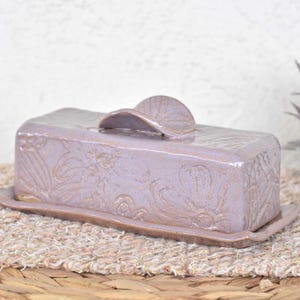 Puede incluir: Una mantequera de cerámica lavanda con tapa y base a juego. El plato presenta patrones florales en relieve y un asa curva en la tapa. El plato rectangular se asienta sobre una estera tejida.