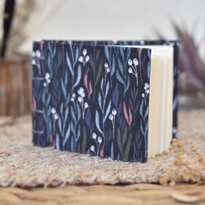 May include: A small, open journal with a dark blue cover featuring a botanical pattern of white berries, blue and green leaves, and pink accents. The pages are cream-colored and visible. The journal is resting on a woven surface.