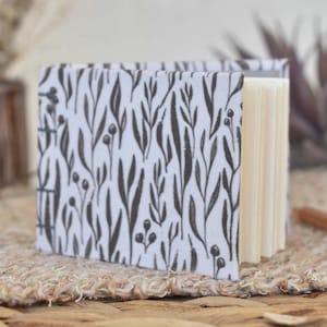 May include: A small, open notebook with a white cover featuring a black botanical pattern of leaves and berries. The pages are a light cream color. The notebook is sitting on a woven surface.