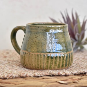 May include: A white ceramic mug with a black handle and the text "But First, Coffee" in a black, bold font. The mug is sitting on a wooden surface. The mug is approximately 4 inches tall.