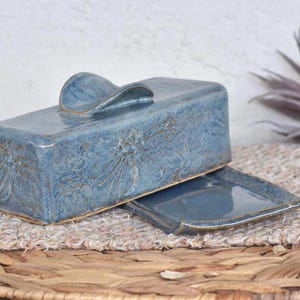 Könnte beinhalten: Rechteckige blaue Keramik-Butterdose mit Deckel und Griff. Die Dose hat einen passenden Boden und ist mit einem floralen Muster verziert. Die Butterdose steht auf einer gewebten Matte. Maße in Zentimetern.