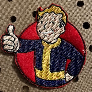 May include: Embroidered patch featuring a cartoon character with blonde hair, a blue shirt, and a yellow vest. The character is giving a thumbs up and is set against a red background.