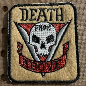 May include: Embroidered patch with the words "DEATH FROM ABOVE" in black lettering. The patch features a white skull with a red and silver design on a tan background, with a black border.