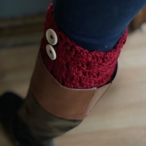 May include: A close-up of a burgundy crochet boot cuff with two white buttons. The cuff is worn over a dark blue garment and a brown and black boot. The crochet has a textured appearance.