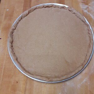 K&ouml;nnte beinhalten: Ein runder, brauner Pizzateig in einer silbernen Form auf einer Holzoberfl&auml;che.