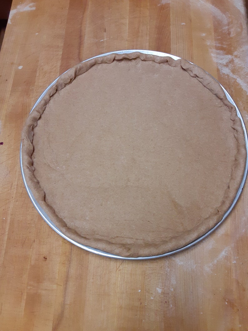 K&ouml;nnte beinhalten: Ein runder, brauner Pizzateig in einer silbernen Form auf einer Holzoberfl&auml;che.