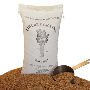 May include: A canvas bag of Liberty Grains Hard Red Winter Wheat Berries, net weight 5 lbs (2.27 kg), with a bundle of wheat stalks graphic. A brass scoop rests in a pile of wheat berries. Text on the bag reads "Since 1950".
