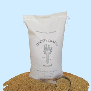 Puede incluir: Una bolsa de lona blanca llena de granos de trigo blanco suave. La bolsa tiene el texto "LIBERTY GRAINS SINCE 1950" impreso en ella. La bolsa está sentada sobre una pila de granos de trigo. Una taza medidora llena de granos de trigo está en primer plano.