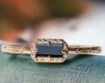 Puede incluir: Un anillo de oro con una piedra rectangular negra engastada en un marco adornado y detallado. El anillo está sobre una superficie azul texturizada.