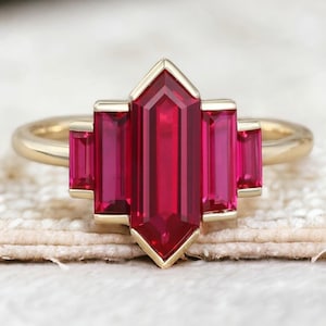 Könnte beinhalten: Ein goldener Ring mit einem großen, tief roten, smaragdgeschliffenen Rubin, umgeben von drei kleineren, baguettegeschliffenen Rubinen.