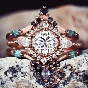 May include: A rose gold engagement ring with a large round center diamond surrounded by smaller diamonds and black gemstones. The ring has a delicate, intricate design with a bohemian aesthetic.