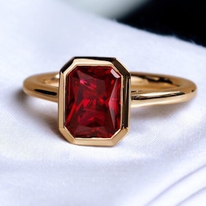 以下が含まれることがあります： 大きな長方形の深い赤色の宝石をあしらったゴールドリング。 宝石は金のベゼルにセットされ、リングバンドはシンプルな磨かれた金です。 リングは白い布の背景に表示されています。