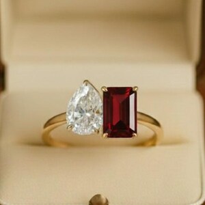 Può includere: Un anello d'oro con due pietre preziose: una pietra bianca a forma di pera e una pietra rossa rettangolare. L'anello è presentato in un cofanetto in similpelle marrone con interno color crema.