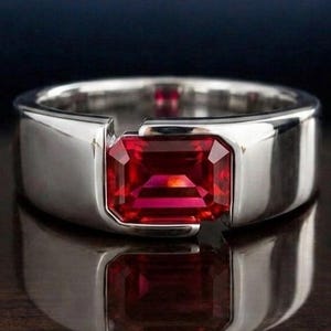 May include: A silver ring featuring a rectangular, bright red gemstone. The ring has a modern, minimalist design with a polished finish. The gemstone is set in a bezel setting, creating a sleek and elegant look. The ring is displayed on a reflective surface.