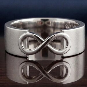 Può includere: Un anello d'argento con un simbolo dell'infinito. L'anello ha una fascia larga e piatta con finitura lucida. Il simbolo dell'infinito è in rilievo e centrale. L'anello è mostrato su una superficie riflettente.