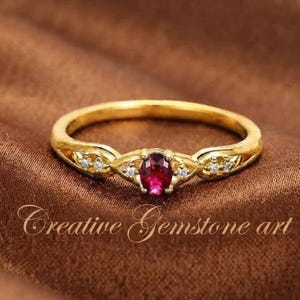 May include: A gold ring featuring a central oval-cut red gemstone flanked by two small clear stones on each side. The band has a polished finish. The text Creative Gemstone art is visible at the bottom of the image.