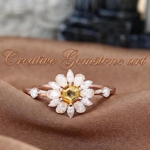 Puede incluir: Un anillo de oro rosa con una piedra preciosa amarilla hexagonal central rodeada por un halo de piedras blancas de talla marquesa dispuestas como pétalos de flor. El texto Creative Gemstone art es visible sobre el anillo de banda fina.