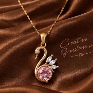 May include: A gold-toned necklace featuring a swan-shaped pendant. The pendant is set with a round pink gemstone at the base, three marquise-cut clear stones forming the wing, and a small clear stone at the eye. Text reads Creative Gemstone art.